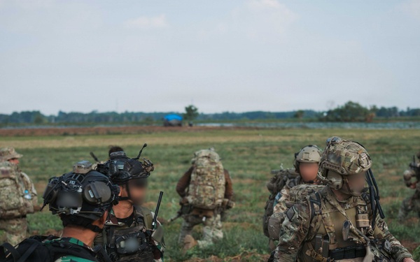 The image shows a joint military exercise or operation involving soldiers from at least two nations in an open field environment. A soldier in the foreground wears an Indonesian flag patch (red and white) on green camouflage uniform, while others wear MultiCam/OCP pattern uniforms consistent with Western military forces. Faces have been digitally blurred, suggesting operational security considerations.