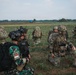 The image shows a joint military exercise or operation involving soldiers from at least two nations in an open field environment. A soldier in the foreground wears an Indonesian flag patch (red and white) on green camouflage uniform, while others wear MultiCam/OCP pattern uniforms consistent with Western military forces. Faces have been digitally blurred, suggesting operational security considerations.
