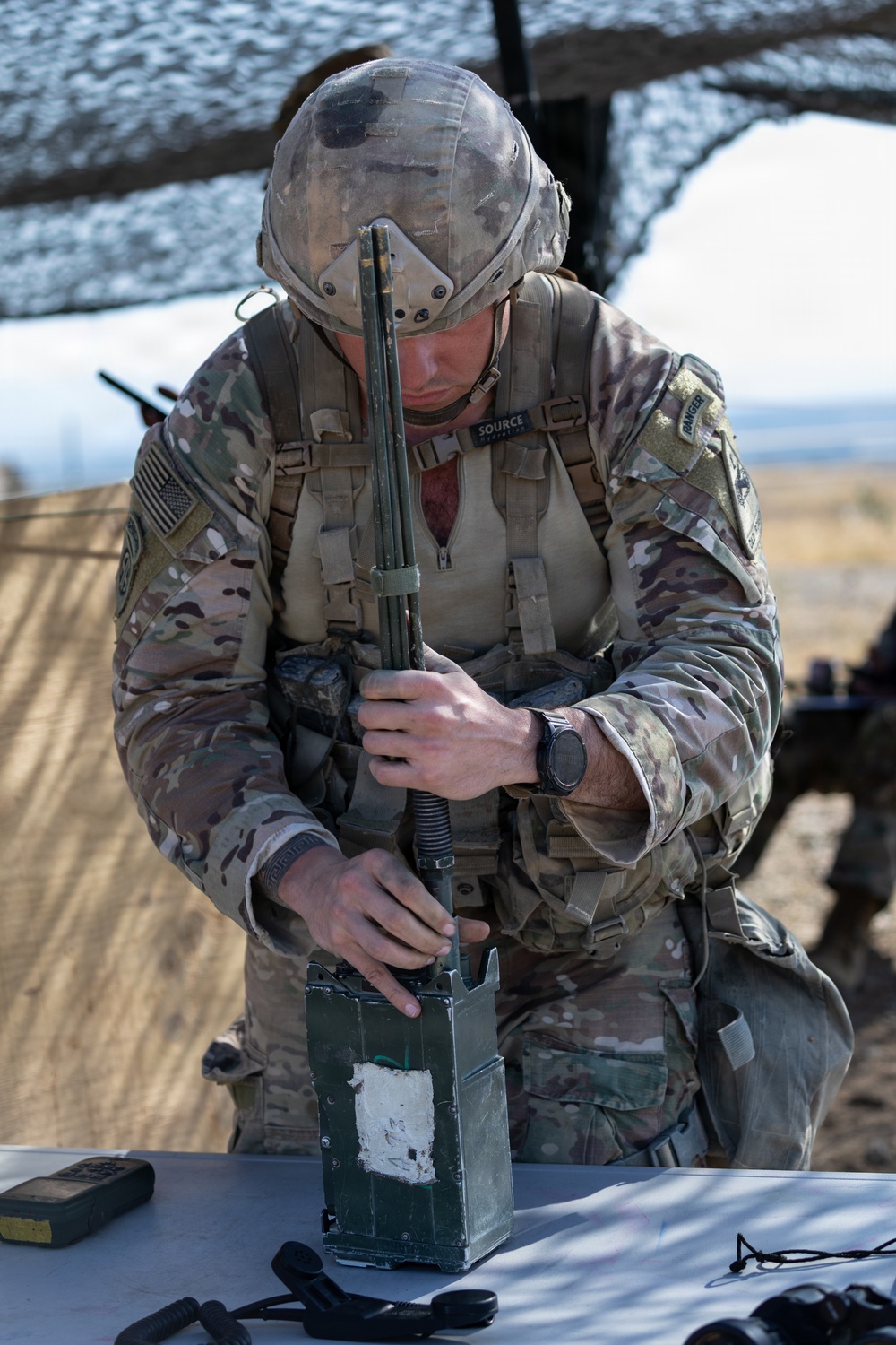 A U.S. Army soldier in full MultiCam (OCP) combat uniform and helmet is working with a military radio system at a field operations table under camouflage netting. The soldier appears to be assembling or configuring a radio handset with an antenna, connecting it to a base unit. A 'RANGER' tab is visible on the left shoulder, indicating the soldier has completed Ranger School.