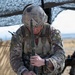 A U.S. Army soldier in full MultiCam (OCP) combat uniform and helmet is working with a military radio system at a field operations table under camouflage netting. The soldier appears to be assembling or configuring a radio handset with an antenna, connecting it to a base unit. A 'RANGER' tab is visible on the left shoulder, indicating the soldier has completed Ranger School.