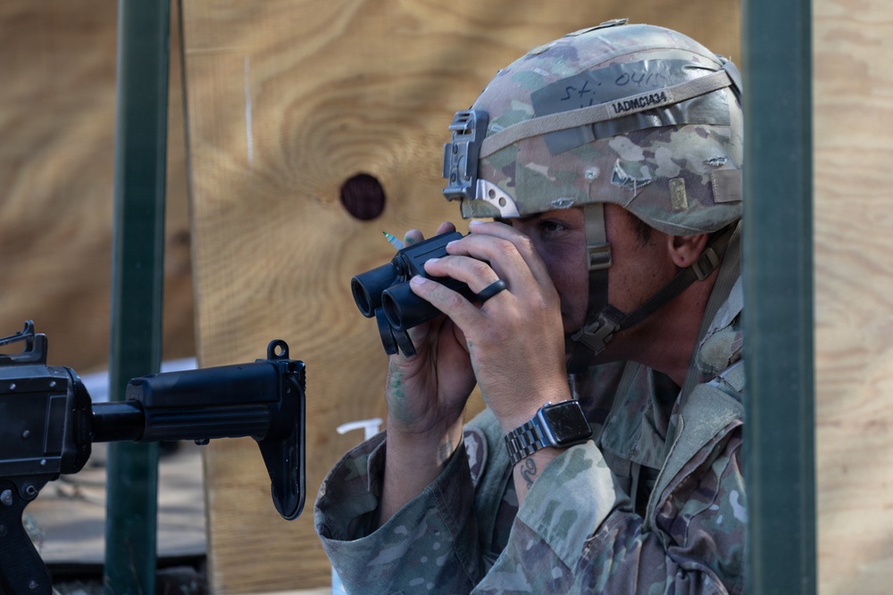 A soldier in MultiCam/OCP camouflage uniform and FAST-style helmet is using a compact rangefinder or monocular device while positioned near a rifle. A plywood target board with a dark circular aiming point is visible in the background. The scene appears to be a military training range or qualification exercise.