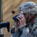 A soldier in MultiCam/OCP camouflage uniform and FAST-style helmet is using a compact rangefinder or monocular device while positioned near a rifle. A plywood target board with a dark circular aiming point is visible in the background. The scene appears to be a military training range or qualification exercise.