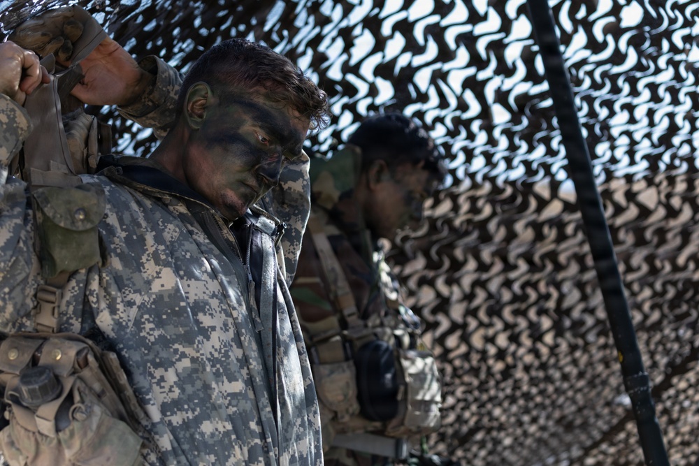 The image shows military personnel in full combat gear with camouflage face paint, standing beneath a camouflage netting structure. At least two soldiers are visible, with the foreground soldier wearing US Army ACU (Army Combat Uniform) digital camouflage pattern. Their heads are bowed, suggesting a moment of prayer, reflection, or a briefing.
