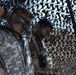 The image shows military personnel in full combat gear with camouflage face paint, standing beneath a camouflage netting structure. At least two soldiers are visible, with the foreground soldier wearing US Army ACU (Army Combat Uniform) digital camouflage pattern. Their heads are bowed, suggesting a moment of prayer, reflection, or a briefing.