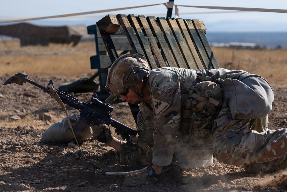 A soldier in full combat gear, including a helmet and body armor, is crouched low to the ground handling an assault rifle, likely an M4 carbine. The setting appears to be a dry, arid training range with wooden pallet barriers and sandbags visible in the background. The soldier appears to be conducting a tactical drill or weapons handling exercise.