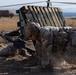 A soldier in full combat gear, including a helmet and body armor, is crouched low to the ground handling an assault rifle, likely an M4 carbine. The setting appears to be a dry, arid training range with wooden pallet barriers and sandbags visible in the background. The soldier appears to be conducting a tactical drill or weapons handling exercise.