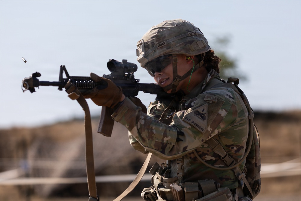 A U.S. Army soldier in full combat gear is actively firing an M4 carbine rifle, with a spent brass casing visibly ejecting from the weapon. The soldier is wearing MultiCam (OCP) camouflage uniform with a tactical helmet, body armor, and hearing protection. Unit patches visible on the uniform include 'Old Ironsides' (1st Armored Division) designation.
