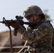 A U.S. Army soldier in full combat gear is actively firing an M4 carbine rifle, with a spent brass casing visibly ejecting from the weapon. The soldier is wearing MultiCam (OCP) camouflage uniform with a tactical helmet, body armor, and hearing protection. Unit patches visible on the uniform include 'Old Ironsides' (1st Armored Division) designation.