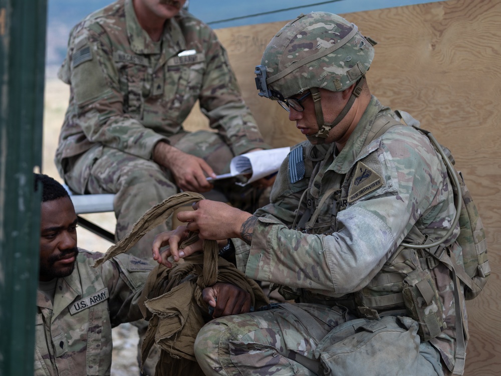 Three U.S. Army soldiers in Operational Camouflage Pattern (OCP) uniforms are engaged in what appears to be a field training exercise or medical/equipment drill. One soldier wearing a helmet and glasses is crouching and working with tactical gear or fabric on another seated soldier, while a third soldier sits in the background holding papers, likely observing or evaluating. The setting appears to be an outdoor field environment near a plywood structure.