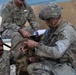 Three U.S. Army soldiers in Operational Camouflage Pattern (OCP) uniforms are engaged in what appears to be a field training exercise or medical/equipment drill. One soldier wearing a helmet and glasses is crouching and working with tactical gear or fabric on another seated soldier, while a third soldier sits in the background holding papers, likely observing or evaluating. The setting appears to be an outdoor field environment near a plywood structure.