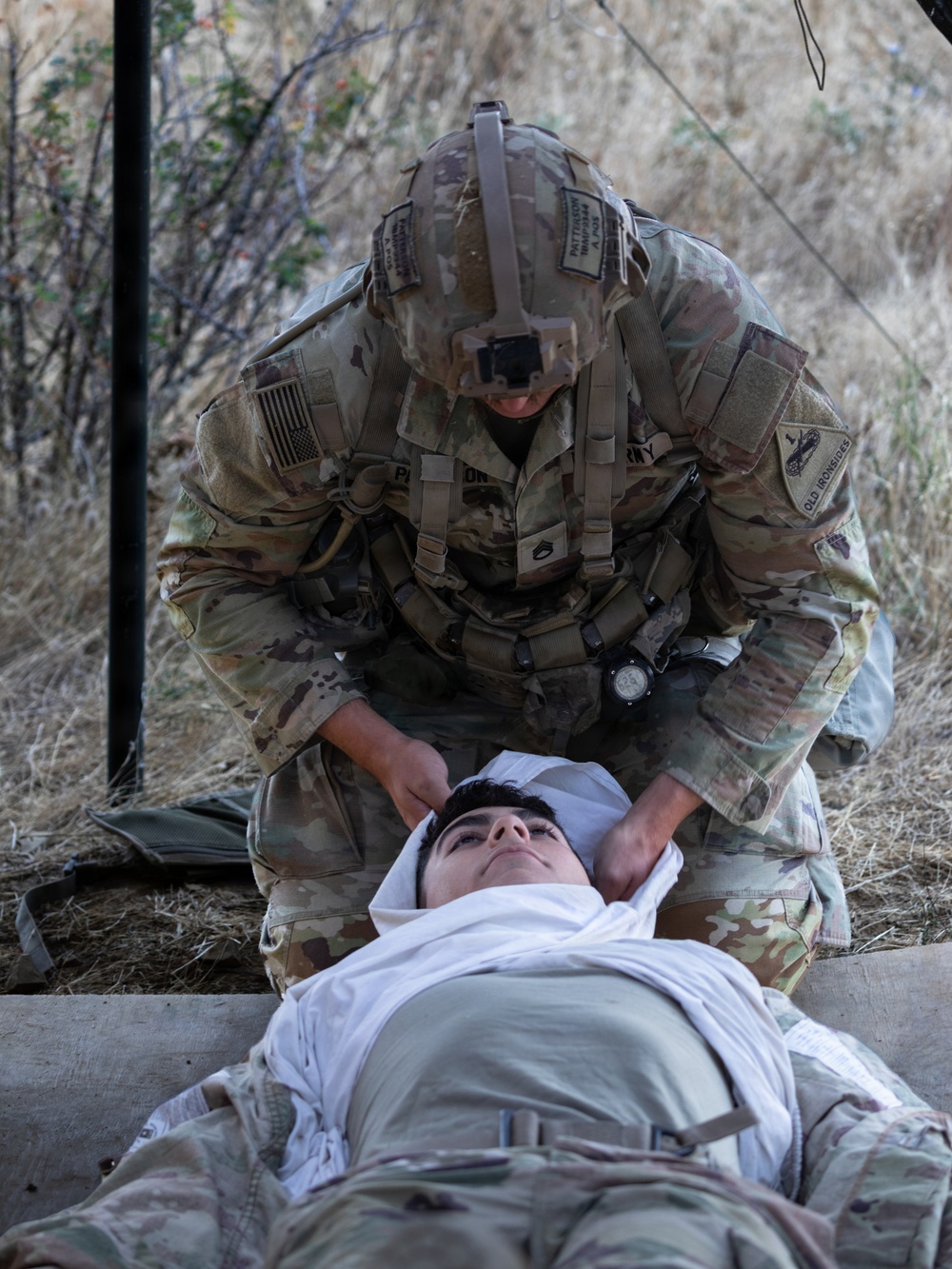 A U.S. Army soldier in full combat gear and OCP (Operational Camouflage Pattern) uniform is performing a medical assessment or stabilization technique on a casualty lying on a litter/stretcher. The soldier appears to be applying a cervical spine or head stabilization hold, suggesting a tactical combat casualty care (TCCC) scenario. The setting appears to be an outdoor field training exercise in a dry, brushy terrain.