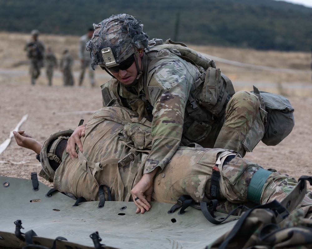 A U.S. Army soldier in full combat gear and OCP (Operational Camouflage Pattern) uniform is performing a casualty evacuation drill, loading or positioning a simulated casualty onto a litter/stretcher. The soldier is wearing a helmet with netting and accessories, sunglasses, and a full tactical kit. Additional soldiers are visible in the background on what appears to be a training range or field exercise area.