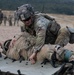 A U.S. Army soldier in full combat gear and OCP (Operational Camouflage Pattern) uniform is performing a casualty evacuation drill, loading or positioning a simulated casualty onto a litter/stretcher. The soldier is wearing a helmet with netting and accessories, sunglasses, and a full tactical kit. Additional soldiers are visible in the background on what appears to be a training range or field exercise area.