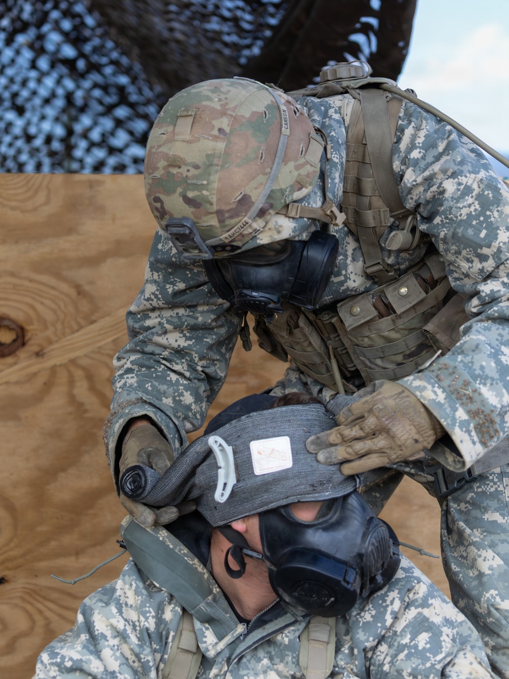 Two military personnel in U.S. Army uniforms are engaged in what appears to be a CBRN (Chemical, Biological, Radiological, Nuclear) training exercise. The standing soldier, wearing a multicam helmet and gas mask, is assisting a seated soldier by fitting or adjusting a protective hood and gas mask. Both are wearing Army Combat Uniforms (ACU) with full protective gear.