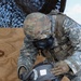 Two military personnel in U.S. Army uniforms are engaged in what appears to be a CBRN (Chemical, Biological, Radiological, Nuclear) training exercise. The standing soldier, wearing a multicam helmet and gas mask, is assisting a seated soldier by fitting or adjusting a protective hood and gas mask. Both are wearing Army Combat Uniforms (ACU) with full protective gear.