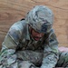 A U.S. Army soldier wearing OCP (Operational Camouflage Pattern) uniform with '1st Armored Division - Old Ironsides' patch is performing tactical combat casualty care (TCCC) training on a medical mannequin. The soldier is applying what appears to be a wound dressing or hemostatic bandage to the mannequin's leg/groin area. The name tape visible reads 'HOWELL' and the soldier wears a patrol cap with a mounted device.