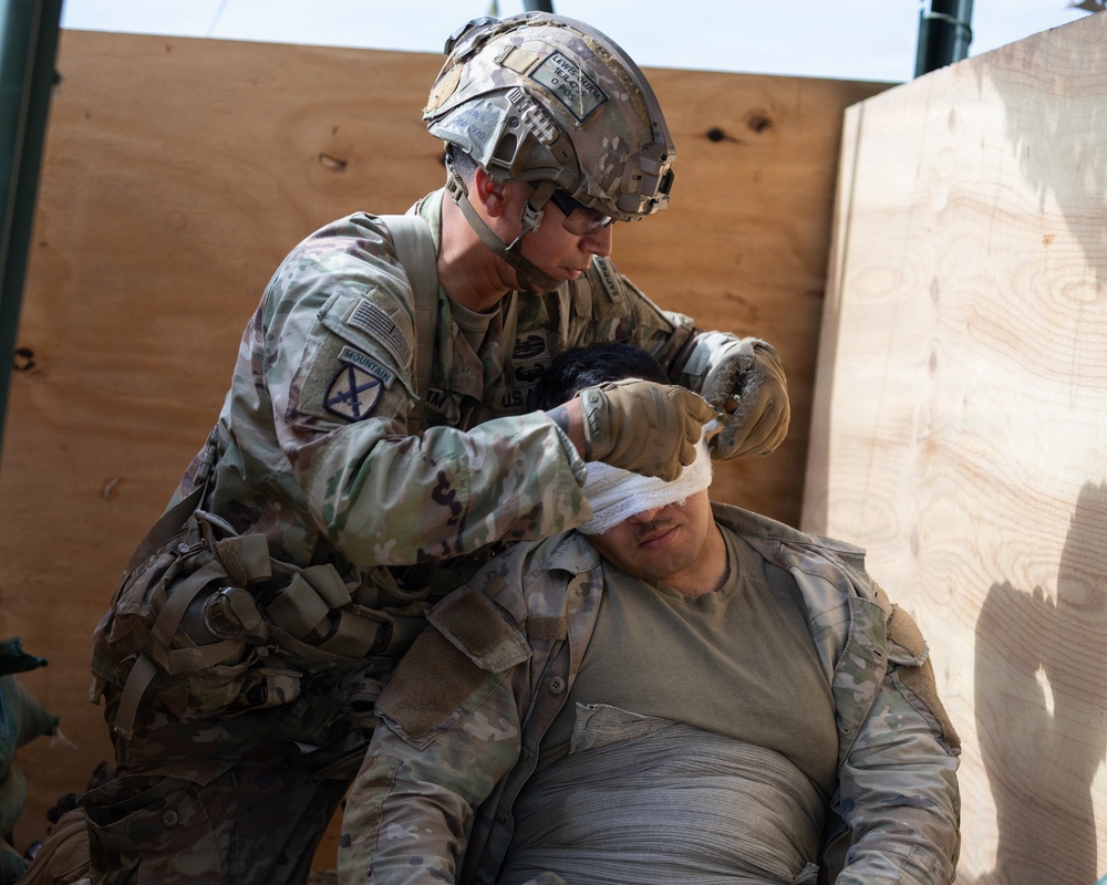 A U.S. Army soldier from the 10th Mountain Division is applying a bandage or dressing to the head/eye area of a casualty during what appears to be a combat casualty care training exercise or real medical treatment. The treating soldier is in full combat gear including a helmet and multicam uniform, while the casualty is leaning against a plywood barrier. The scene depicts tactical combat casualty care (TCCC) procedures being performed.