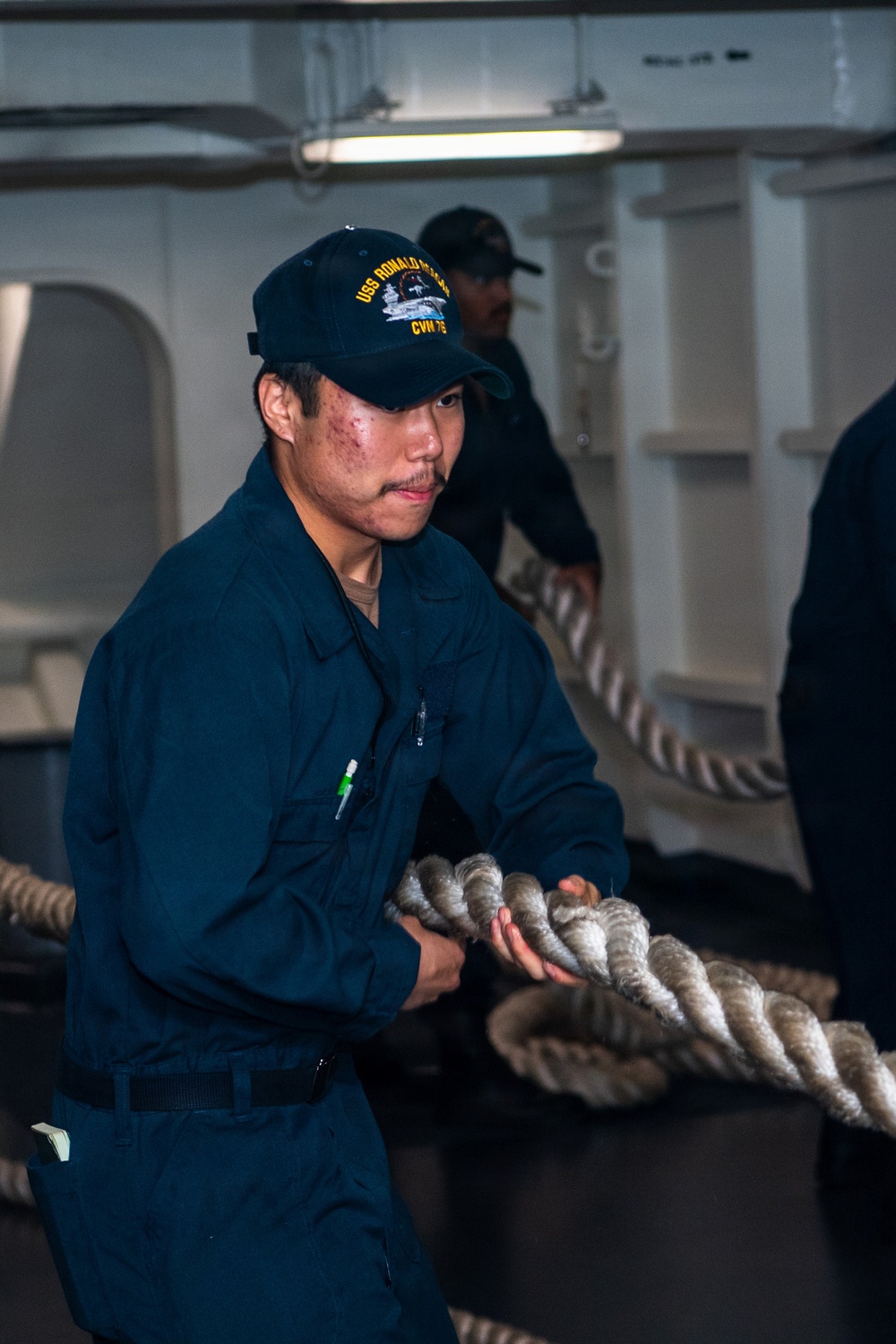 A U.S. Navy sailor aboard the USS Ronald Reagan (CVN-76) is actively handling a large mooring line, pulling or managing the heavy rope during what appears to be a line-handling evolution. He is wearing standard Navy blue coveralls and a ship-specific ball cap with 'USS Ronald Reagan CVN-76' text. A second sailor is visible in the background performing a similar task.