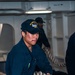 A U.S. Navy sailor aboard the USS Ronald Reagan (CVN-76) is actively handling a large mooring line, pulling or managing the heavy rope during what appears to be a line-handling evolution. He is wearing standard Navy blue coveralls and a ship-specific ball cap with 'USS Ronald Reagan CVN-76' text. A second sailor is visible in the background performing a similar task.