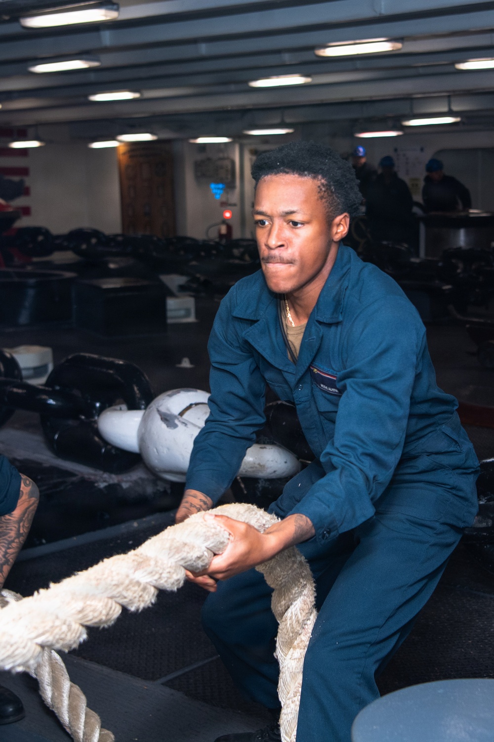 A sailor in dark blue Navy coveralls is pulling a large mooring line aboard what appears to be a naval vessel's interior deck or hangar bay. The individual is exerting significant physical effort, leaning back with both hands gripping a thick white rope. An American flag and other personnel are visible in the background.