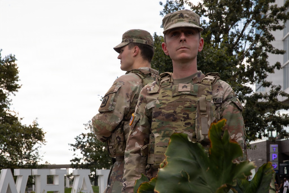 Two U.S. Army soldiers in Operational Camouflage Pattern (OCP) uniforms are standing at attention in an outdoor urban setting. The foreground soldier, wearing a plate carrier vest and bearing Sergeant rank insignia, faces the camera directly while the second soldier stands with their back turned. Both wear the American flag patch on their right shoulder sleeve.