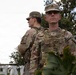 Two U.S. Army soldiers in Operational Camouflage Pattern (OCP) uniforms are standing at attention in an outdoor urban setting. The foreground soldier, wearing a plate carrier vest and bearing Sergeant rank insignia, faces the camera directly while the second soldier stands with their back turned. Both wear the American flag patch on their right shoulder sleeve.