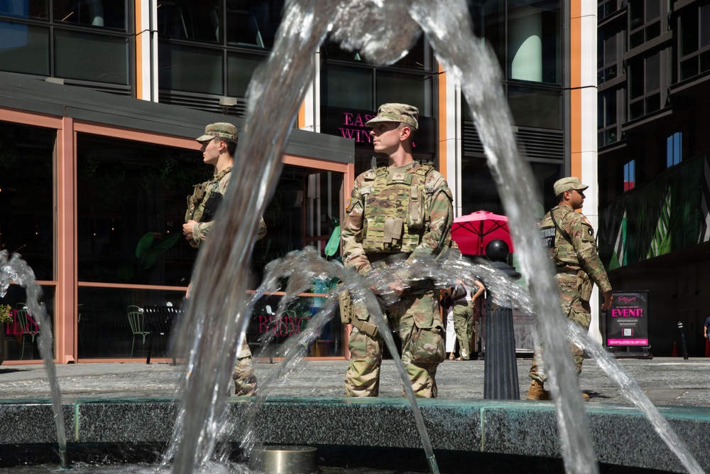 Three soldiers in military camouflage uniforms and patrol caps are standing in what appears to be an urban plaza or commercial area. They are positioned near a decorative water fountain, with a modern building featuring retail or restaurant spaces visible in the background. The soldiers appear to be on guard duty or conducting a security presence mission.