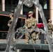 Three soldiers in military camouflage uniforms and patrol caps are standing in what appears to be an urban plaza or commercial area. They are positioned near a decorative water fountain, with a modern building featuring retail or restaurant spaces visible in the background. The soldiers appear to be on guard duty or conducting a security presence mission.