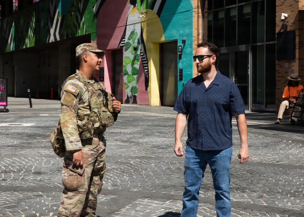 Two men are conversing in an outdoor urban plaza with a colorful mural-covered building in the background. One man is wearing U.S. Army combat uniform (OCP/MultiCam) with a tactical vest and American flag patch, while the other is dressed in civilian clothing with sunglasses. The setting appears to be a modern pedestrian area with cobblestone paving during daytime.