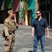 Two men are conversing in an outdoor urban plaza with a colorful mural-covered building in the background. One man is wearing U.S. Army combat uniform (OCP/MultiCam) with a tactical vest and American flag patch, while the other is dressed in civilian clothing with sunglasses. The setting appears to be a modern pedestrian area with cobblestone paving during daytime.