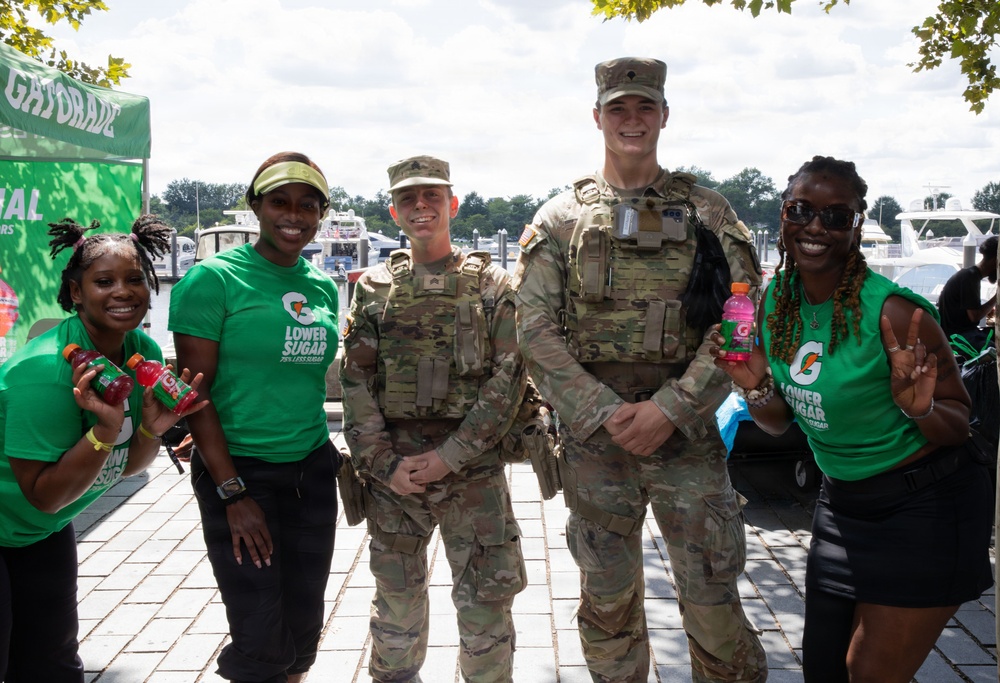Five individuals pose together outdoors near a waterfront area at what appears to be a promotional event. Two military personnel in camouflage uniforms and tactical vests stand alongside three Gatorade brand ambassadors wearing green 'Lower Sugar' promotional t-shirts. The group is smiling and holding Gatorade bottles near a branded Gatorade tent.
