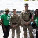 Five individuals pose together outdoors near a waterfront area at what appears to be a promotional event. Two military personnel in camouflage uniforms and tactical vests stand alongside three Gatorade brand ambassadors wearing green 'Lower Sugar' promotional t-shirts. The group is smiling and holding Gatorade bottles near a branded Gatorade tent.
