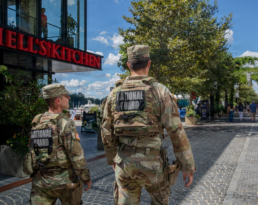 Two U.S. National Guard soldiers in full camouflage uniforms and tactical vests are patrolling a pedestrian waterfront area. Both soldiers have 'NATIONAL GUARD' patches clearly visible on their backs. The setting appears to be a commercial/dining district near a waterway, with a 'Hell's Kitchen' restaurant visible in the background.
