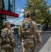 Two U.S. National Guard soldiers in full camouflage uniforms and tactical vests are patrolling a pedestrian waterfront area. Both soldiers have 'NATIONAL GUARD' patches clearly visible on their backs. The setting appears to be a commercial/dining district near a waterway, with a 'Hell's Kitchen' restaurant visible in the background.
