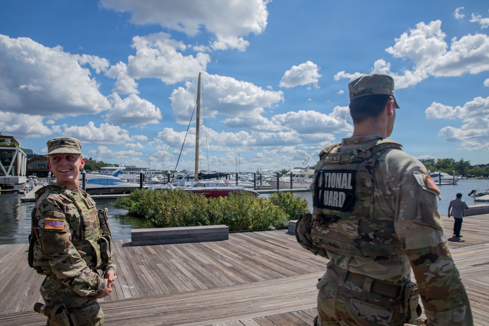 Two U.S. National Guard soldiers in OCP (Operational Camouflage Pattern) uniforms are standing on a wooden boardwalk near a marina. The soldier facing the camera is smiling, while the other has a clearly visible 'NATIONAL GUARD' patch on their back. The setting appears to be a civilian waterfront area with numerous boats and yachts docked in the background.