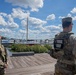 Two U.S. National Guard soldiers in OCP (Operational Camouflage Pattern) uniforms are standing on a wooden boardwalk near a marina. The soldier facing the camera is smiling, while the other has a clearly visible 'NATIONAL GUARD' patch on their back. The setting appears to be a civilian waterfront area with numerous boats and yachts docked in the background.