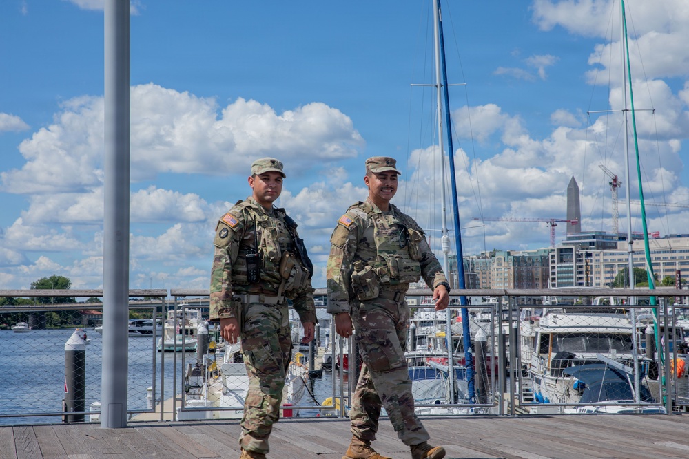 Two U.S. Army soldiers in OCP (Operational Camouflage Pattern) uniforms are walking along a waterfront boardwalk, appearing to conduct a patrol. In the background, a marina with multiple boats and the Washington Monument are clearly visible, indicating the location is Washington, D.C., likely near the Southwest Waterfront or The Wharf. Both soldiers are wearing tactical vests and appear to be on an active security detail.