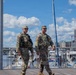 Two U.S. Army soldiers in OCP (Operational Camouflage Pattern) uniforms are walking along a waterfront boardwalk, appearing to conduct a patrol. In the background, a marina with multiple boats and the Washington Monument are clearly visible, indicating the location is Washington, D.C., likely near the Southwest Waterfront or The Wharf. Both soldiers are wearing tactical vests and appear to be on an active security detail.
