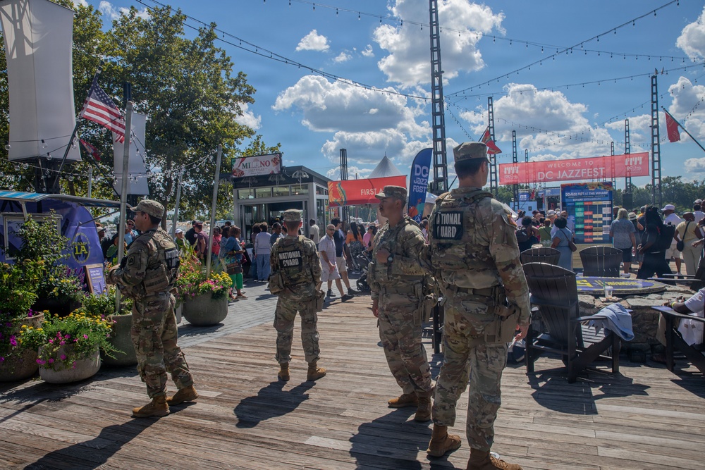 National Guard personnel are stationed at the DC JazzFest event at The Wharf in Washington, D.C., providing security presence at the outdoor festival. Four uniformed soldiers in camouflage with 'NATIONAL GUARD' patches on their backs are visible on a wooden boardwalk surrounded by festival-goers. The scene depicts a sunny day with crowds, vendor signage, string lights, and festival banners in the background.