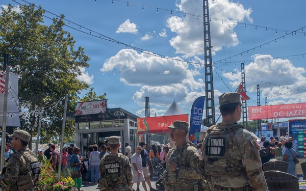National Guard personnel are stationed at the DC JazzFest event at The Wharf in Washington, D.C., providing security presence at the outdoor festival. Four uniformed soldiers in camouflage with 'NATIONAL GUARD' patches on their backs are visible on a wooden boardwalk surrounded by festival-goers. The scene depicts a sunny day with crowds, vendor signage, string lights, and festival banners in the background.