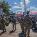 National Guard personnel are stationed at the DC JazzFest event at The Wharf in Washington, D.C., providing security presence at the outdoor festival. Four uniformed soldiers in camouflage with 'NATIONAL GUARD' patches on their backs are visible on a wooden boardwalk surrounded by festival-goers. The scene depicts a sunny day with crowds, vendor signage, string lights, and festival banners in the background.