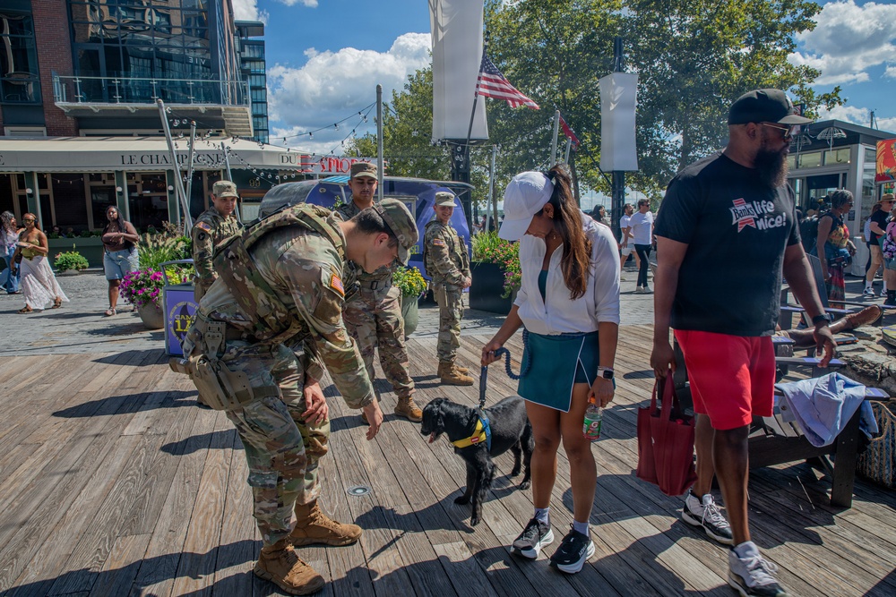 A group of U.S. Army soldiers in OCP (Operational Camouflage Pattern) uniforms interact with civilians on a wooden boardwalk in what appears to be a waterfront public area. One soldier is bending down to pet a small black dog on a leash held by a woman in a white cap and green skirt. The setting appears to be a public event or community engagement activity near a restaurant called 'Le Champagne' with an American flag visible in the background.