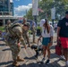 A group of U.S. Army soldiers in OCP (Operational Camouflage Pattern) uniforms interact with civilians on a wooden boardwalk in what appears to be a waterfront public area. One soldier is bending down to pet a small black dog on a leash held by a woman in a white cap and green skirt. The setting appears to be a public event or community engagement activity near a restaurant called 'Le Champagne' with an American flag visible in the background.