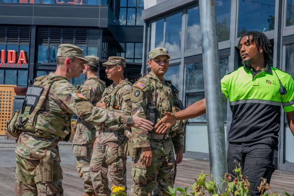 A group of U.S. Army soldiers in OCP (Operational Camouflage Pattern) uniforms are interacting with a civilian security worker wearing a high-visibility green polo shirt outside a commercial building. One soldier and the civilian appear to be shaking hands, suggesting a cordial or professional greeting. The setting appears to be an urban outdoor area, possibly during a community engagement or security coordination event.