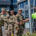 A group of U.S. Army soldiers in OCP (Operational Camouflage Pattern) uniforms are interacting with a civilian security worker wearing a high-visibility green polo shirt outside a commercial building. One soldier and the civilian appear to be shaking hands, suggesting a cordial or professional greeting. The setting appears to be an urban outdoor area, possibly during a community engagement or security coordination event.