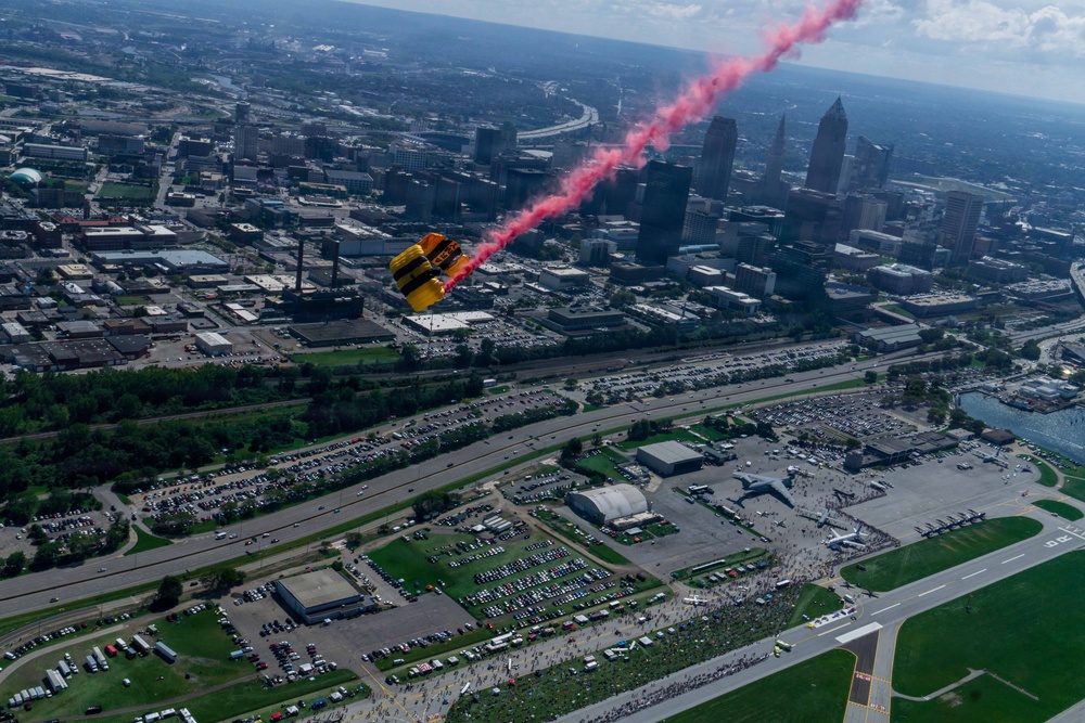 An aerial photograph captures a skydiver with a yellow and black parachute trailing red smoke over a major urban area, with a large airshow venue visible below. The city skyline, identifiable by its distinctive skyscrapers, is visible in the background along a waterfront. Large crowds and parked vehicles are visible around what appears to be an airport or airfield hosting a public airshow event.