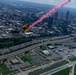 An aerial photograph captures a skydiver with a yellow and black parachute trailing red smoke over a major urban area, with a large airshow venue visible below. The city skyline, identifiable by its distinctive skyscrapers, is visible in the background along a waterfront. Large crowds and parked vehicles are visible around what appears to be an airport or airfield hosting a public airshow event.