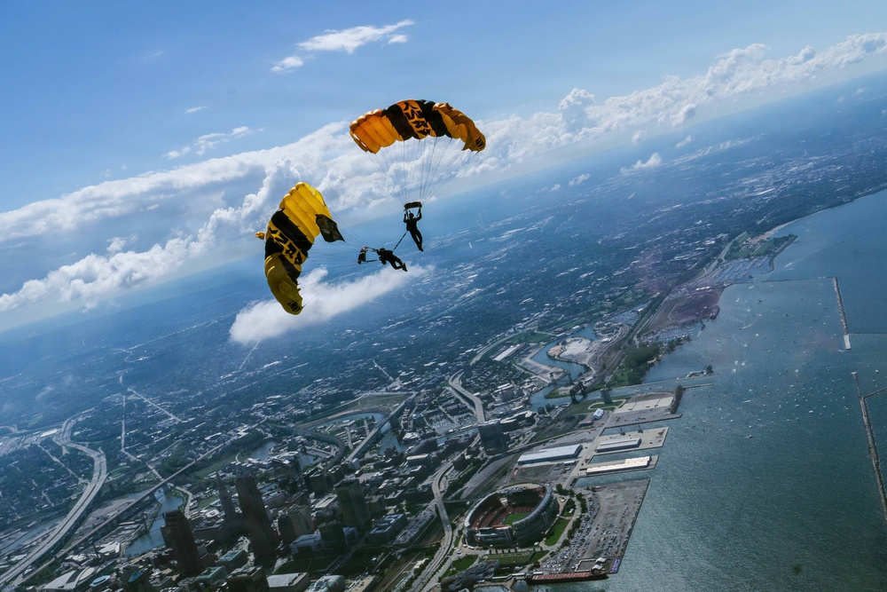 Two skydivers with deployed yellow and black parachutes are descending over a large coastal city, photographed from an aerial perspective. The parachutes display markings consistent with a military or professional demonstration team. The city below features a waterfront, stadium, highway infrastructure, and urban sprawl extending to the horizon.