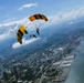 Two skydivers with deployed yellow and black parachutes are descending over a large coastal city, photographed from an aerial perspective. The parachutes display markings consistent with a military or professional demonstration team. The city below features a waterfront, stadium, highway infrastructure, and urban sprawl extending to the horizon.