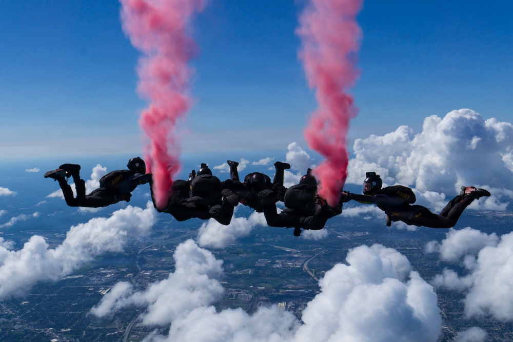 A group of skydivers in black uniforms perform a formation freefall at high altitude, releasing pink/red colored smoke from canisters attached to their gear. The jumpers appear to be members of a military demonstration team, identifiable by 'ARMY' patches visible on their uniforms. They are holding a horizontal linked formation against a backdrop of blue sky, cumulus clouds, and an urban landscape far below.